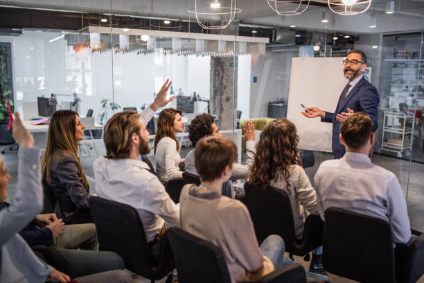 Mature businessman talking to large group of people on a business presentation in a board room. Some of them are raising their hands.